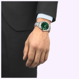 A person in a dark suit and light blue shirt wears a Tissot Watches Gentleman 38mm T1658071109100 with a green dial on their left wrist, set against a plain light background.