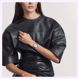 A woman in a black leather top sits against a plain, light-colored background, her left hand touching her chest as she wears the Tissot Watches PR 100 34mm T1502101103100, featuring a stainless steel case and Swiss Quartz movement.