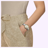 A person wearing beige high-waisted pants with a tied belt has their right hand in their pocket, showcasing the Tissot Watches PR 100 34mm T1502102103100 on their wrist—a stainless steel timepiece by Tissot Watches featuring a white face, gold accents, and precise Swiss Quartz movement.