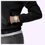 A person dressed in a black jacket and gray jeans stands with their hand in their pocket, showcasing the Tissot Watches PR 100 36mm T1502171111100 by Tissot Watches. The backdrop is a light purple shade.