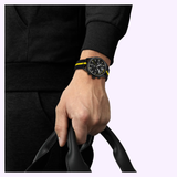 Wearing a bold black and yellow Tissot Watches PR 100 Tour de France 2026 40mm T1504173705700 chronograph by Tissot Watches, an individual in a sleek black long-sleeve outfit holds the handles of a black bag, posed against a light purple background.
