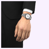 A person in a black suit wears a Tissot Watches PR516 38mm T1494071103100, a Swiss automatic watch by Tissot Watches featuring a white dial and metal bracelet, on their left wrist. Their relaxed hand is visible against a light background.