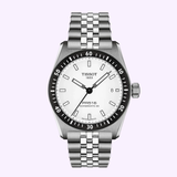 The Tissot Watches PR516 38mm T1494071103100 features a stainless steel case, white dial, black bezel, silver hour markers, date window at 3 o’clock, and a metal link bracelet. Set against a light purple background, this Swiss automatic timepiece is powered by the renowned Powermatic 80 movement.
