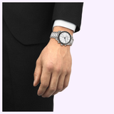 A person in a black suit with a white shirt cuff showcases their left wrist wearing the Tissot Watches PR516 40mm (T1494171101100), featuring a white dial and classic stainless steel bracelet.