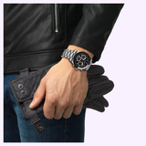 A person in a black leather jacket and jeans holds black gloves in one hand, wearing a Tissot Watches PR516 41mm T1494592105100 by Tissot Watches, featuring a stainless steel case, black dial with multiple subdials, and powered by an intricate Swiss manual winding movement.