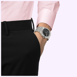 A person wearing black pants and a pink shirt has their hand in their pocket, showcasing the Tissot Watches PRX 41mm T1374171105100 by Tissot Watches with its sleek black dial.