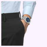 A person dressed in black pants and a light blue shirt has their right hand tucked into their pocket, subtly showcasing a Tissot Watches PRX 41mm T1374171105100—a sophisticated stainless steel timepiece from Tissot Watches that lends an elegant touch to the look.