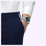 A person wearing navy pants, a white shirt, and a Tissot Watches PRX 42mm T1374271104101 by Tissot Watches, featuring a sapphire crystal and black dial, with their right hand in their pocket against a light background.