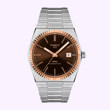 The Tissot Watches PRX Powermatic 80 Steel & 18K Gold Bezel 40mm (T9314074129100) features a brushed silver metal bracelet, brown dial, gold hour markers, date window at 3 o’clock, and an elegant 18K gold bezel.