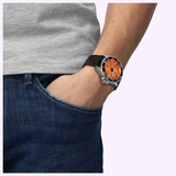A person in blue jeans and a gray t-shirt, with one hand in their pocket, showcases the Tissot Watches Seastar 2000 44mm T1209071728100—a striking Swiss automatic watch with an orange dial and black strap.