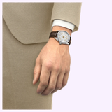 A person in a beige suit with a white shirt cuff showing wears a Tissot Watches Visodate 39mm T1574071603100 Swiss automatic timepiece with a brown leather strap on their left wrist, set against a light purple background.