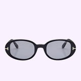 Tom Ford Sunglasses FT1364/s 4901A in a sleek black oval design, showcasing gray lenses and distinctive T-shaped metallic accents at the upper corners of the frames. The sunglasses are set against a solid light purple background, creating a chic Miami-inspired ambiance.