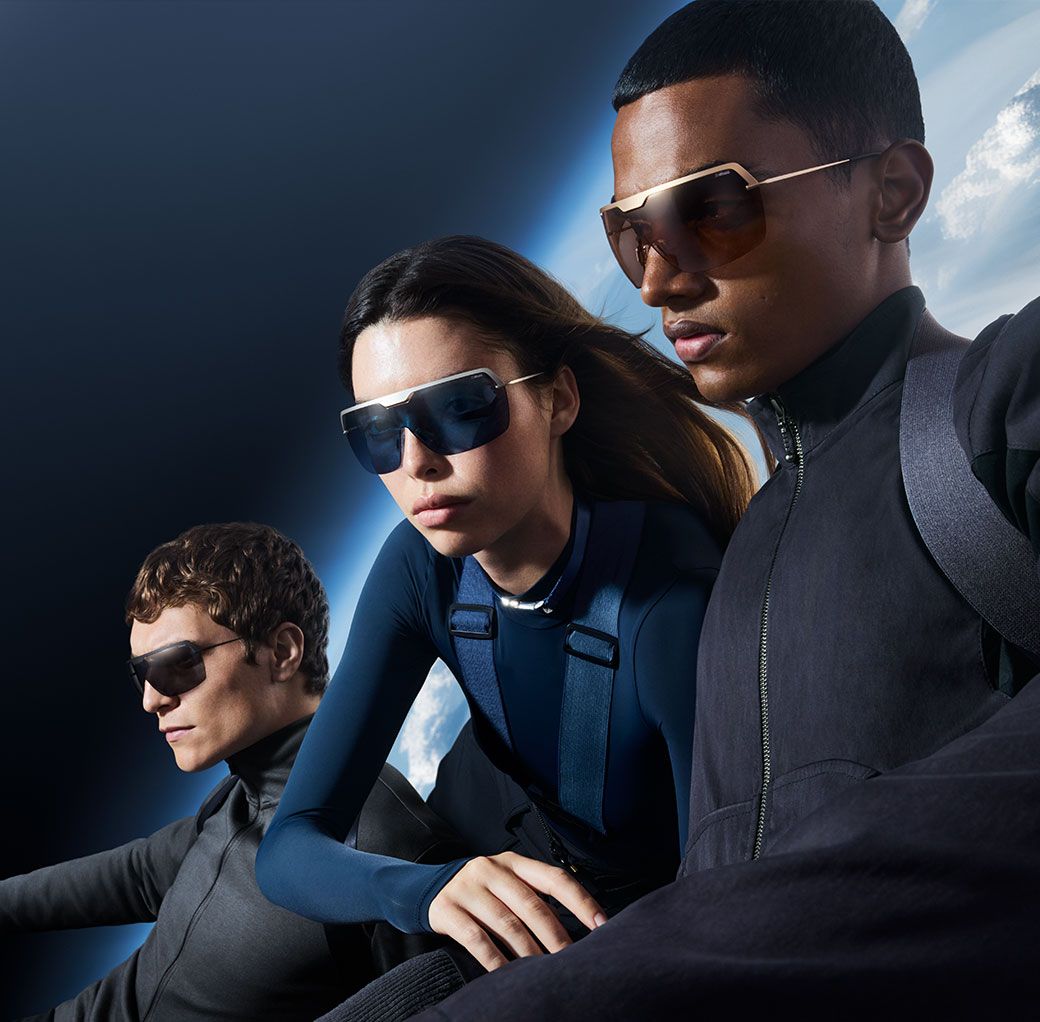 Three people wearing dark, stylish sunglasses and modern, sleek clothing pose confidently against a dramatic, space-like background with clouds and a blue horizon.