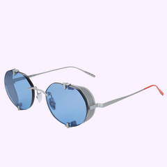 The Vysen Sunglasses The Uttar UT4 Unisex (UPC: 3423104192179) feature a round silver frame, blue-tinted lenses, silver arms with red tips, and side shields. Handcrafted in Italy by Vysen Sunglasses, shown on a pastel purple background.