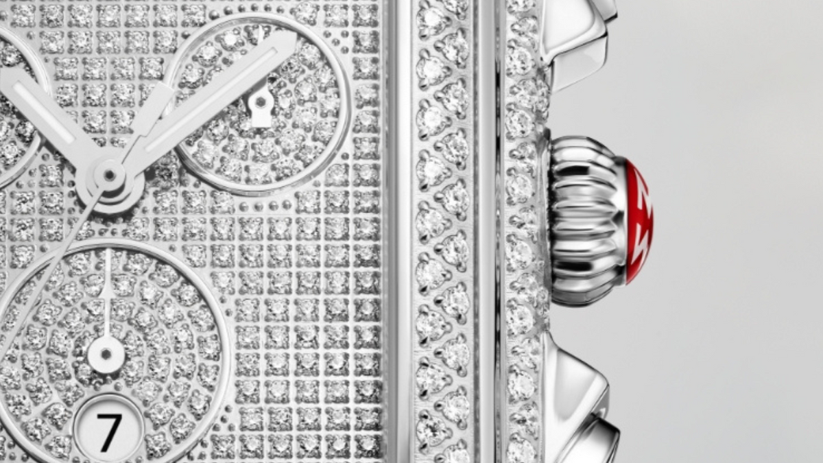 Close-up of a luxury watch face encrusted with diamonds, showing part of the dial with subdials, date window, and a silver crown with a red accent on the side.