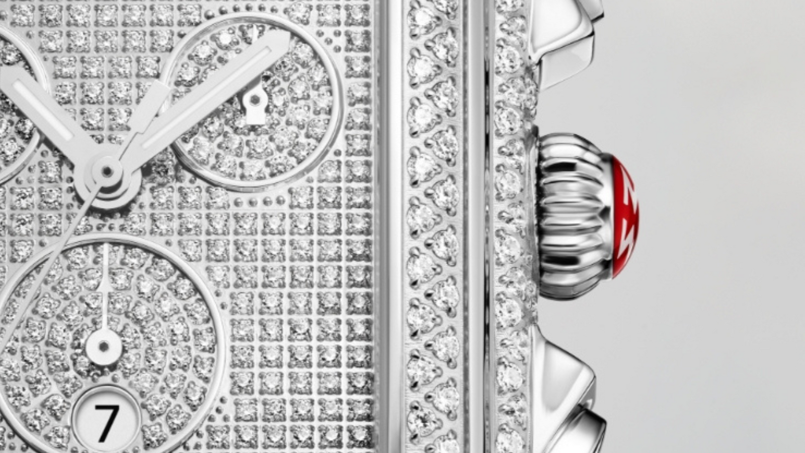 Close-up of a luxury watch face encrusted with diamonds, showing part of the dial with subdials, date window, and a silver crown with a red accent on the side.