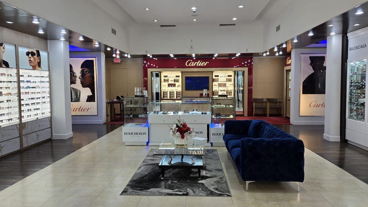 A luxury eyewear store interior featuring displays of sunglasses, branded sections for Cartier and Balenciaga, a blue velvet sofa, a marble coffee table with a flower arrangement, and bright, modern lighting.