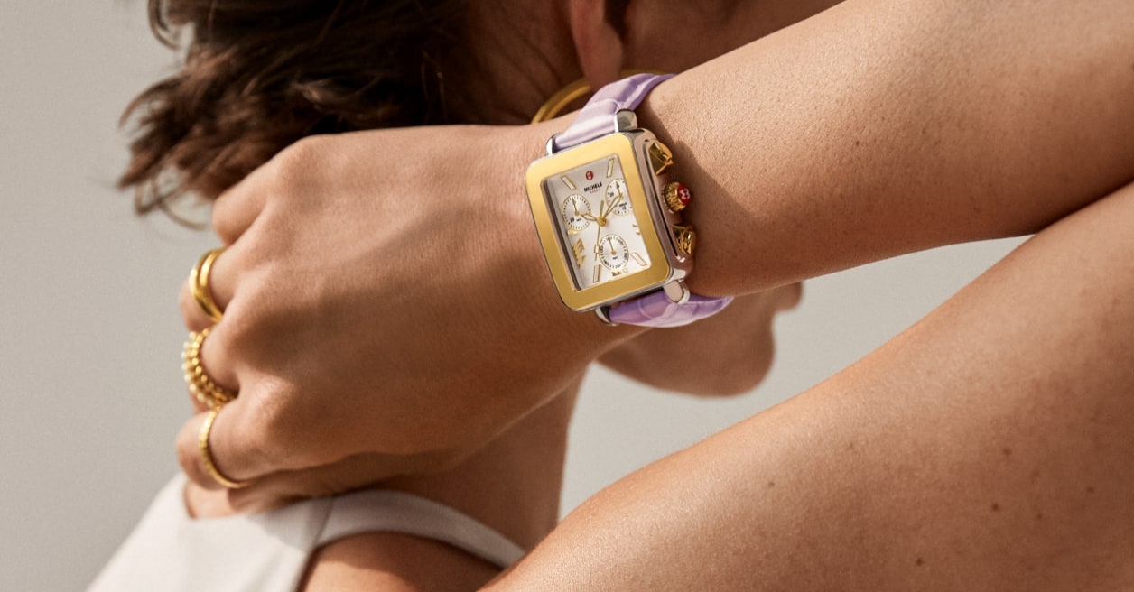 Close-up of a person wearing a square-faced gold smartwatch with a lavender strap, along with several gold rings and a bracelet, against a neutral background.