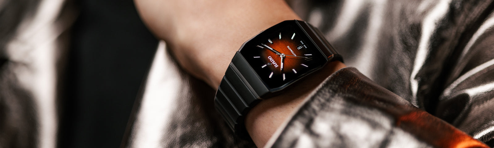 A close-up of a person’s wrist wearing a black smartwatch with an orange digital watch face, dressed in a shiny metallic silver jacket.