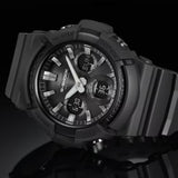 A CASIO G-Shock Watch ANALOG-DIGITAL GAS100B-1A from G-Shock Watches, featuring a black design with a thick bezel, silver hour markers, and side buttons, is displayed elegantly on a reflective black surface.