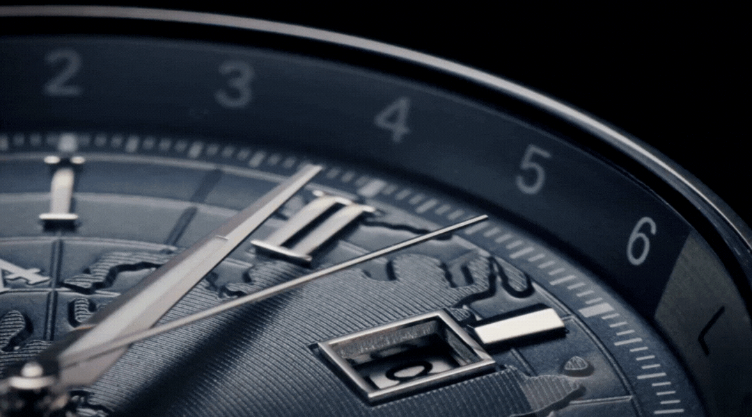 Close-up of a luxury watch face, showing intricate details, including textured world map patterns, silver watch hands, and a date window, with numbers 2 to 6 visible along the dial’s edge.