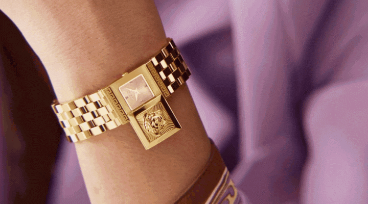 A close-up of a gold wristwatch with a checkered band on a persons wrist. The watch face is partly covered by a hinged lid featuring an engraved lions head. The person is wearing a lavender sleeve.