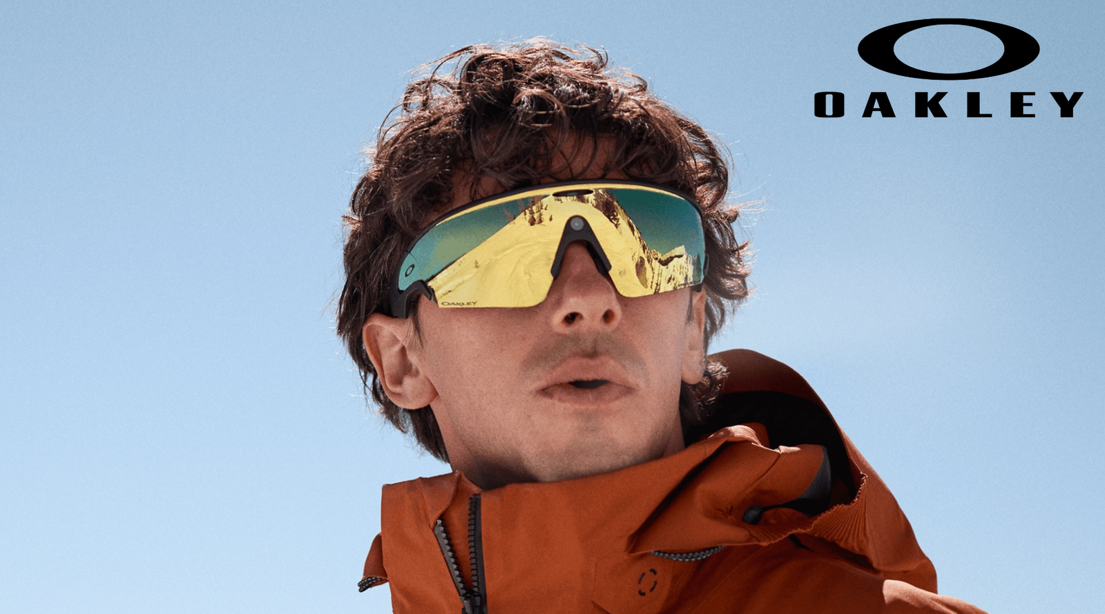 A person in an orange jacket wears reflective Oakley ski goggles against a clear blue sky, with the Oakley logo in the top right corner. Snowy mountains are visible in the reflection of the goggles.