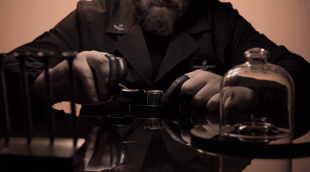 A person in a dark coat uses precision tools to work on a small object at a polished table, with various instruments and a glass dome nearby, under warm lighting.