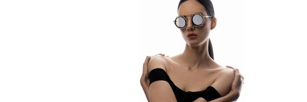 A woman with slicked-back hair wearing round, reflective sunglasses and a black off-shoulder top poses confidently against a white background, arms crossed over her chest.