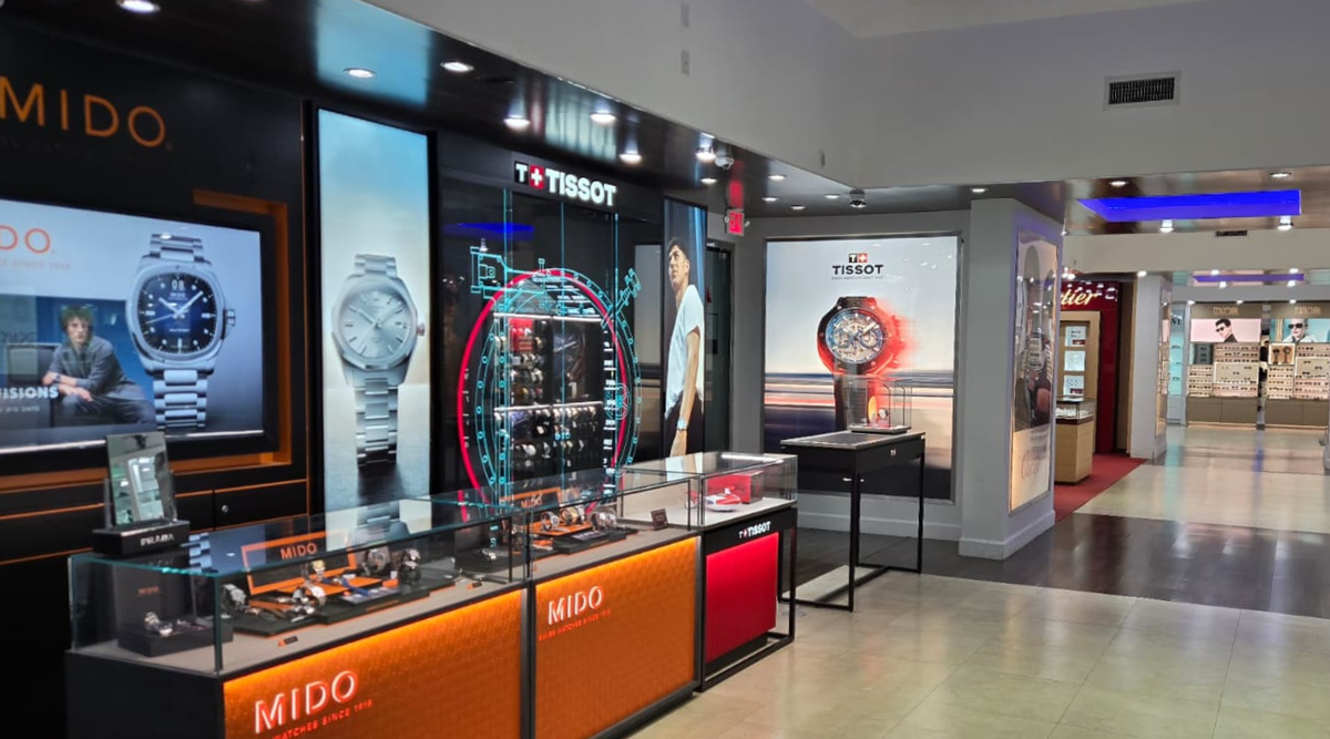 Watch store interior showing Mido and Tissot displays with watches inside glass cases, large branded posters on the walls, and bright lighting illuminating the products.