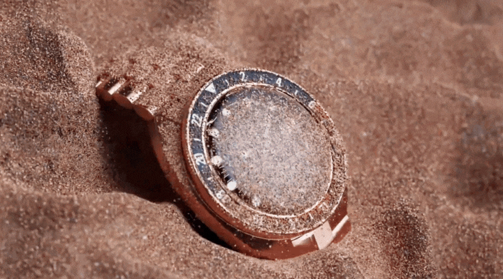 A gold wristwatch partially buried in dry, loose sand, with sand grains covering its face and band. The watch appears weathered, suggesting exposure to outdoor elements.