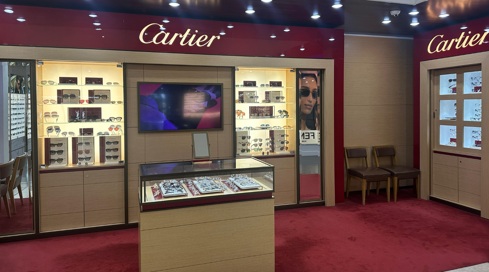 Luxury Cartier eyewear boutique with red carpeting, display cases filled with glasses, a central glass-topped counter, illuminated shelving, wall mirrors, and two chairs in the corner. A screen displays an image on the back wall.