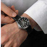 A person in a white shirt adjusts a Bulova Watches Marine Star 98B465 men’s watch by Bulova Watches, featuring a black and silver case with a black strap on their left wrist. The round watch face displays white markers and numbers, highlighting the refined style of this automatic dive watch.