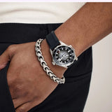 A close-up of a wrist adorned with the Bulova Watches Marine Star 98B465—an elegant silver luxury timepiece with a black strap—paired with a chunky silver chain bracelet. The wearer is dressed in dark pants and a light top, with one hand casually tucked into their pocket.