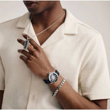 A person wearing a cream textured short-sleeve shirt, a silver pendant necklace, a silver bracelet, and the Bulova Watches Marine Star 98B465 automatic dive watch by Bulova Watches, holding the pendant with their left hand.