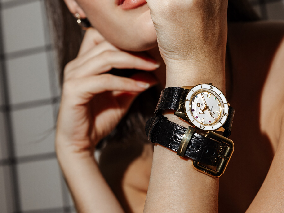 A close-up of a woman resting her chin on her hand, wearing a stylish wristwatch with a white face and a thick black leather strap featuring two gold buckles. The background has a tiled pattern.