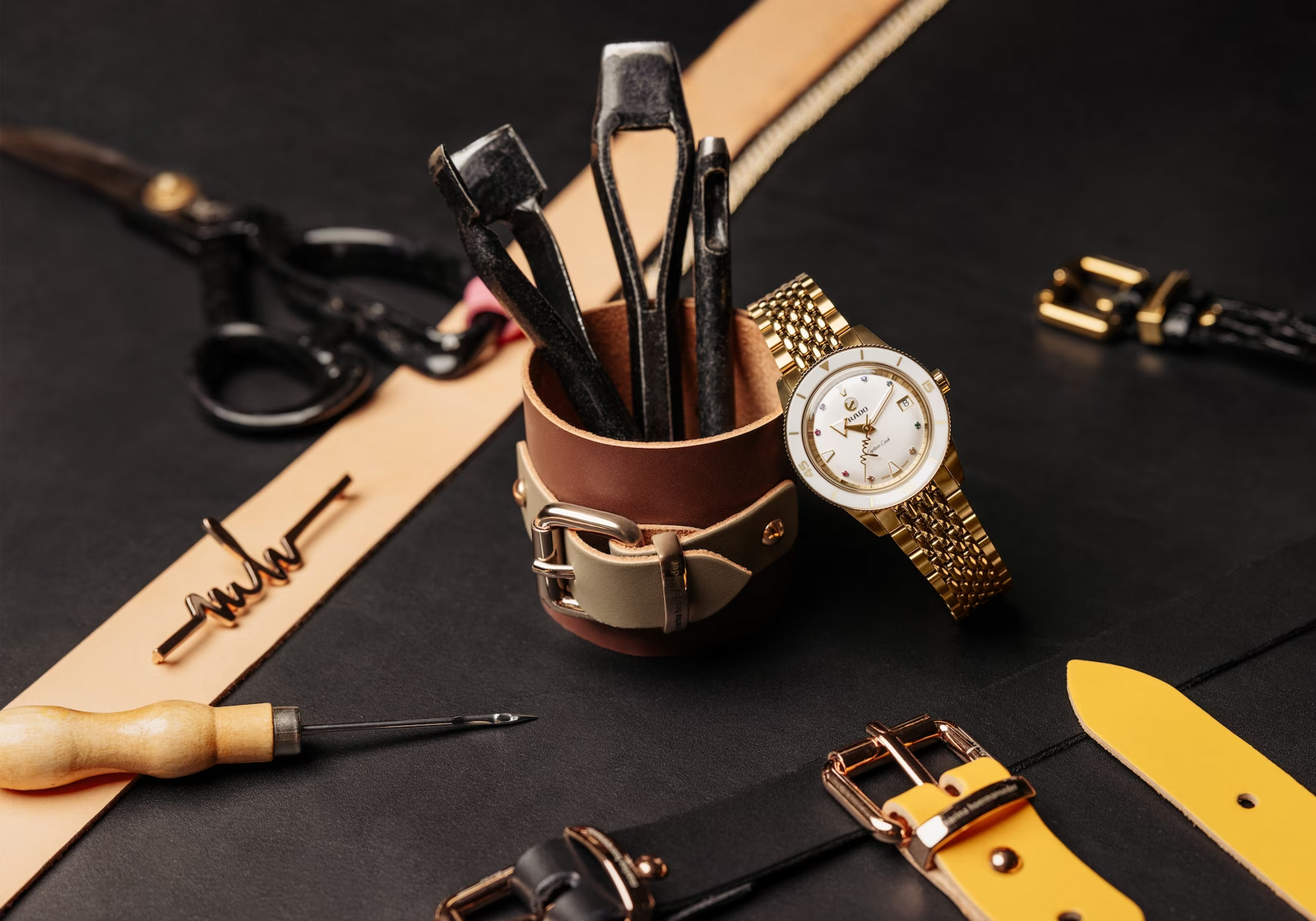 A gold wristwatch rests on a brown leather tool holder filled with metal tools, surrounded by watch straps, buckles, scissors, and leather crafting tools on a dark surface.