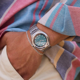 Wearing a Citizen Watches Zenshin Three-hand AW0130-85X men’s watch by Citizen Watches, which features Eco-Drive movement, a silver band, and a turquoise face, along with a thin silver bracelet, the person is dressed in a colorful striped shirt and cream pants.