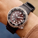 A close-up of the Citizen Watches Promaster Dive E365 BN1024-01Z by Citizen Watches, featuring a black strap on a person’s wrist. This professional diver watch showcases a black and silver bezel and a striking red-black gradient dial with large, luminous markers.