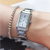 A Citizen Watches Citizen L Bianca EW5620-55A women’s luxury watch, featuring a silver rectangular face with Roman numerals and powered by Eco-Drive technology, is worn on the wrist beside a beaded bracelet, highlighting its elegant design.