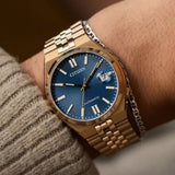 A Citizen Watches TSUYOSA 60 NK0023-57L automatic watch, featuring a gold-tone case and band with a striking blue sunray dial and date window, is worn on a person’s wrist alongside a silver chain bracelet and the beige sleeve of a sweater.