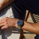 A person wearing the Citizen Watches TSUYOSA Shore NJ0230-59L, featuring a silver and blue design with a blue sunray dial, sits at a table holding a folded newspaper. The white tabletop and natural sunlight emphasize the elegance of this Citizen Watches timepiece on their wrist.
