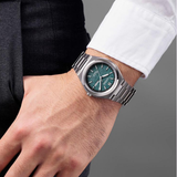 Wearing a white shirt and dark pants, a person showcases the Citizen Watches Zenshin Three-hand AW0130-85X by Citizen Watches—a silver men’s watch with a green textured face and metal band, featuring Eco-Drive movement—on their left wrist against a plain grey background.
