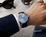 A man in a blue suit checks the time on his Seiko Luxe Watches Presage Cocktail Time Man SRPB43, featuring a light blue face and black leather strap. Sunglasses, a drink, notebook, and pen sit on the marble table—style fit for Miami.
