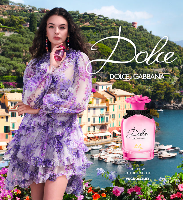 A woman in a frilly, purple floral dress holds a pink perfume bottle. She stands by a scenic seaside village with colorful buildings. The text reads: Dolce & Gabbana Dolce the new eau de toilette #DGDolceLily.