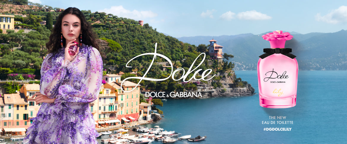 A woman in a flowing purple floral dress stands beside a scenic coastal village. Next to her is a pink Dolce & Gabbana Dolce perfume bottle with a flower cap. Text reads: Dolce & Gabbana, the new eau de toilette, #DOLCELILLY.