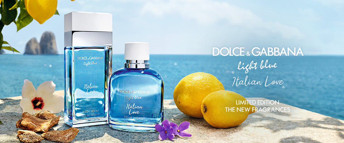 Two bottles of Dolce & Gabbana Light Blue Italian Love fragrance sit on a stone ledge beside lemons and purple flowers, with a scenic ocean view and cliffs in the background. The text highlights the limited edition fragrances.
