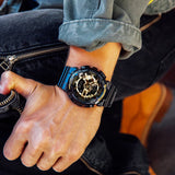 A person dons a green jacket and jeans while seated, holding their knee, with the focus on their wrist wearing the shock-resistant CASIO G-Shock Watch ANALOG-DIGITAL GA110GB-1A by G-Shock Watches in striking black and gold.