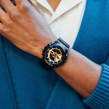 A person in a blue sweater wears a G-Shock Watches CASIO G-Shock Watch ANALOG-DIGITAL GA110GB-1A on their left wrist, with their hand resting on their chest over a white collared shirt.