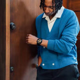 A person with braided hair, dressed in a blue cardigan layered over a white shirt and paired with black pants, stands beside a wooden door. They look down thoughtfully, one hand in their pocket while the other rests on the door. On their wrist is the CASIO G-Shock Watch ANALOG-DIGITAL GA110GB-1A from G-Shock Watches, its durable shock-resistant design adding an understated complement to their relaxed yet stylish appearance.